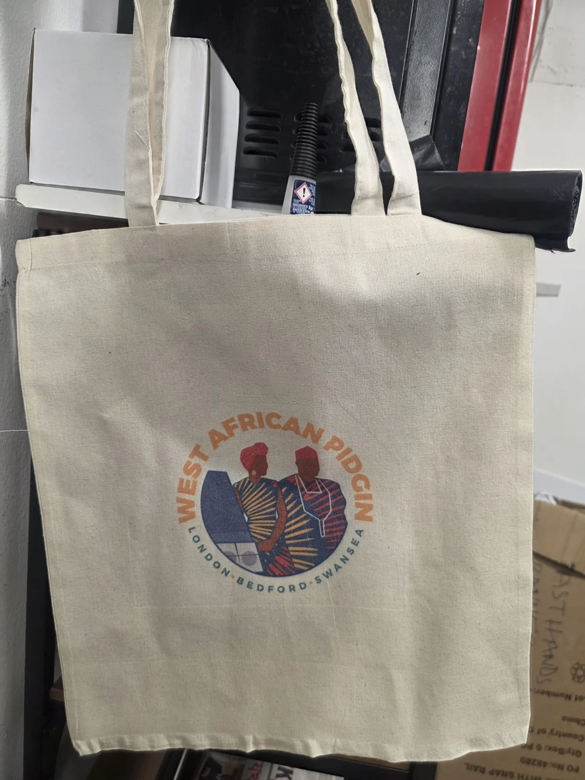 Bulk run of natural cotton tote bags printed with the West African Pidgin logo in full colour — real customer order by IT Wheel London
